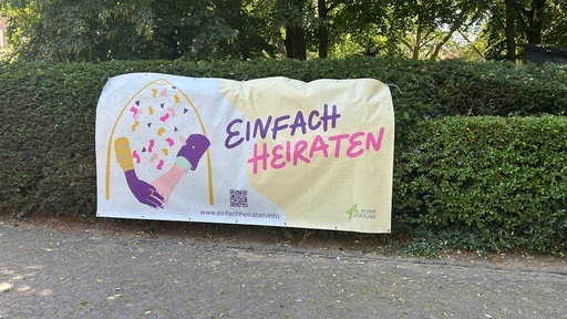 Ein Banner mit der Aufschrift "Einfach Heiraten" ist an einer grünen Hecke befestigt.