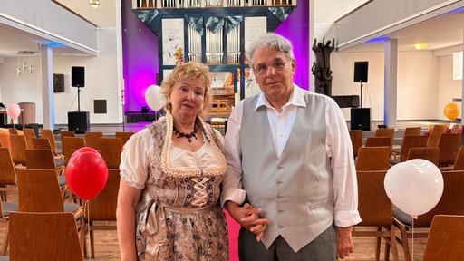 In einer Tracht und Weste heiratet ein älteres Pärchen. Der Raum ist festlich geschmückt.
