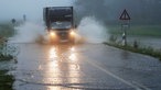 Ein schwarzer Transporter fährt über eine teils überflutete Landstraße, rechts und links von ihm gehen Wasserfontänen ab