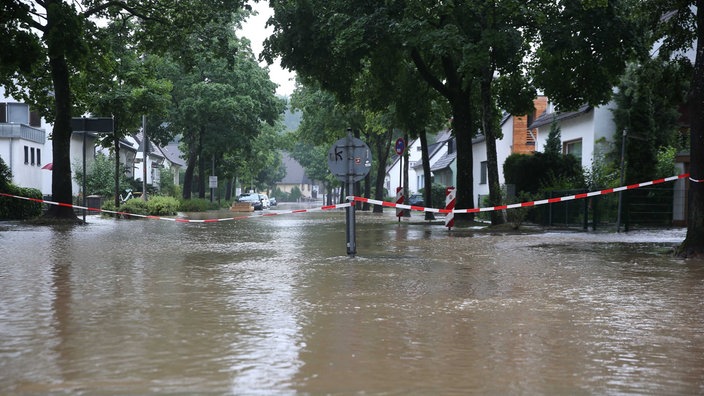 Eine überflutete Straße in Erkrath ist in der Mitte mit einem rot-weißen Flatterband abgesperrt.