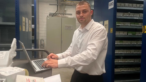 Taras Rudyi steht im weißen Hemd im Lager. Um ihn herum stehen viele Regale, vor ihm sein Laptop.