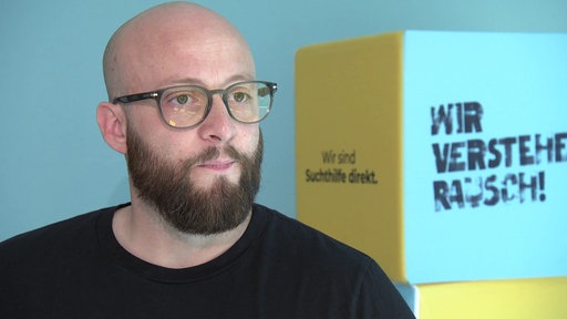 ein Mann in einem schwarzen T-Shirt und Brille steht vor einem gelben Würfel