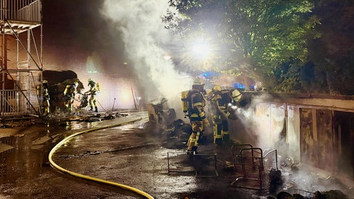 Einsatzkräfte bei Nacht untersuchen qualmende Mülltonnen.