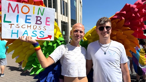 Zwei Teilnehmende der RuhrPride posieren mit großen regenbogenfarbenen Flügeln. Eine Person hält ein Schild mit der Aufschrift "Moin Liebe, tschüss Hass".