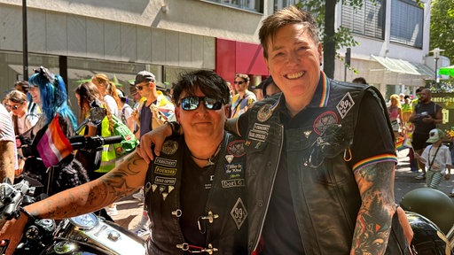 Zwei Personen in schwarzen Lederwesten stehen neben einem Motorrad mit Regenbogenflagge, während im Hintergrund Teilnehmende des RuhrPride durch die Essener Innenstadt ziehen.