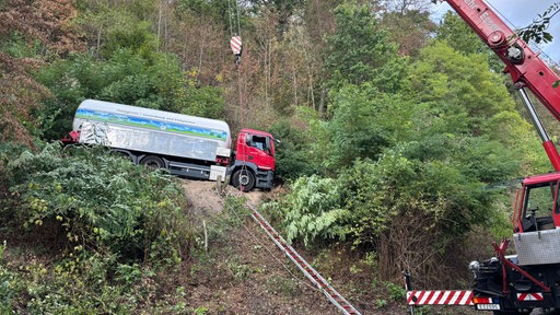 Ein Kran sichert einen roten Tanklaster, der auf einem Abhang feststeckt und geborgen werden soll.