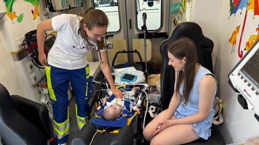 Ein Kind liegt in einem Krankenwagen mit vielen Schläuchen und medizinischen Geräten. Drumherum stehen Mutter und Ärztin.