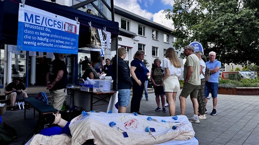 Vor der Essener Uniklinik steht ein Zelt mit einem Schild, auf dem ME/CFS zu lesen ist, davor eine Puppe in einem Bett und Teilnehmer der Mahnwache