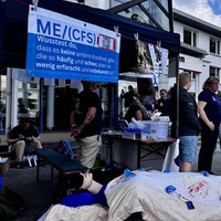 Vor der Essener Uniklinik steht ein Zelt mit einem Schild, auf dem ME/CFS zu lesen ist, davor eine Puppe in einem Bett und Teilnehmer der Mahnwache