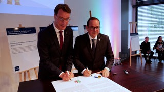 Hendik Wüst und Thomas Kufen stehen vor einem Tisch und unterschreiben ein großes Dokument.