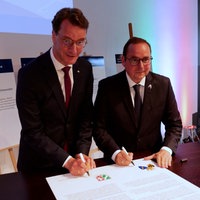 Hendik Wüst und Thomas Kufen stehen vor einem Tisch und unterschreiben ein großes Dokument.