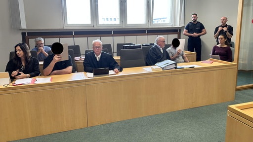 In einem Saal des Essener Landgerichts sitzen die beiden Angeklagten mit vor das Gesicht gehaltenen Papieren und ihre Verteidiger auf der Anklagebank