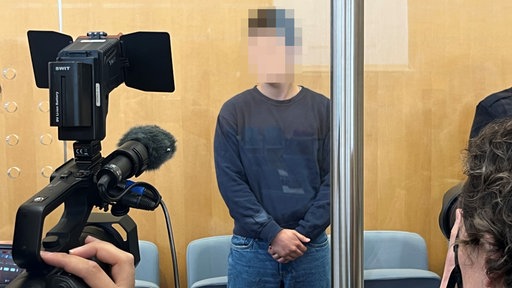 Das Bild zeigt den Angeklagten im Gerichtssaal, wo er von einem Fernsehteam gefilmt wird.