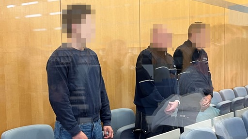 Das Bild zeigt den Angeklagten neben zwei Sicherheitsbediensteten.