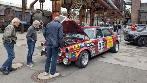 Drei Männer stehen vor einem roten Mercedes-Benz 300 SEL auf einem Industriegelände, die Motorhaube ist geöffnet, im Hintergrund weitere Mercedes-Autos