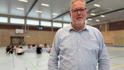 Schulleiter Lukas Rüenauver schaut in der Sporthalle lächelnd in die Kamera 