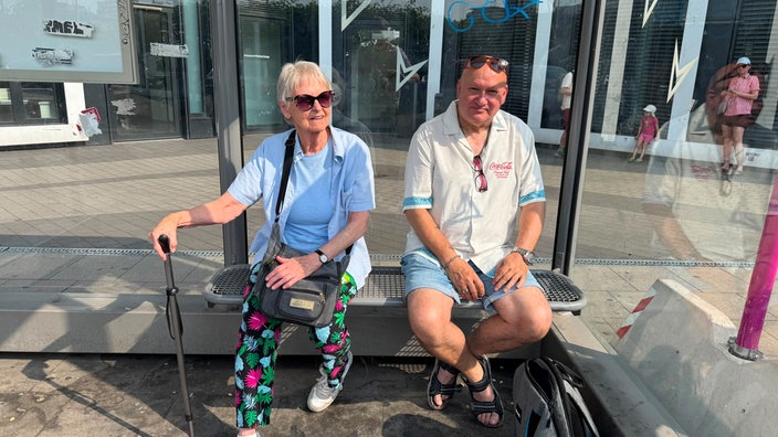 Ein Mann und eine Frau sitzen mit Koffer an einer Haltestelle 