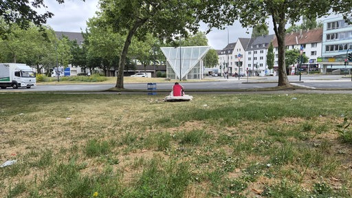 Ein Mann sitzt mit dem Rücken zur Kamera allein auf einer Wiese, dahinter fährt ein LKW eine Hauptstraße entlang