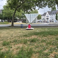 Ein Mann sitzt mit dem Rücken zur Kamera allein auf einer Wiese, dahinter fährt ein LKW eine Hauptstraße entlang