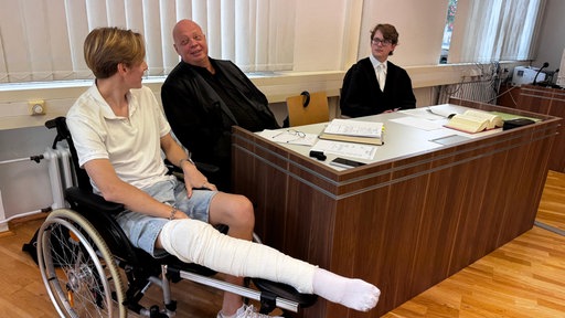Ein junger Mann im Rollstuhl mit Beingips und zwei Männer in schwarzen Roben sitzen vor einem Holzpult