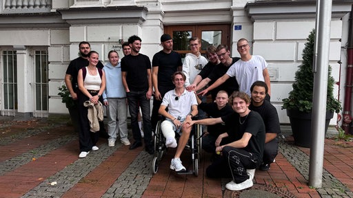 In der Bildmitte ist ein junger Mann im Rollstuhl, um ihn herum stehen und hocken 11 junge Männer und 2 junge Frauen