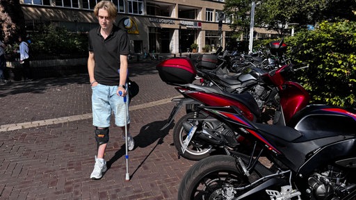 Junger Mann an einer Krücke überquert Platz, im Vordergrund Motorräder