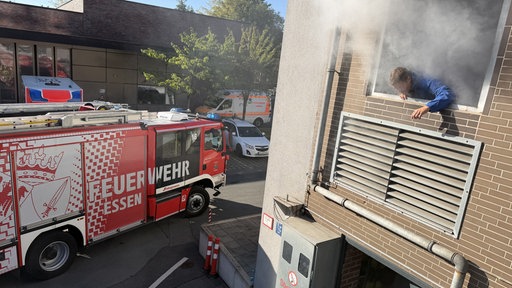 Eine Person neigt sich aus einem qualmenden Fenster im ersten Stock, davor steht ein Feuerwehrfahrzeug.