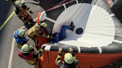 Eine Person liegt in einem Sprungpolster umringt von Feuerwehrleuten.