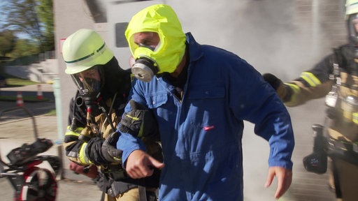 Zwei Feuerwehrleute halten führen einen Betroffenen in Brandfluchtmaske aus einem rauchenenden Gebäude.