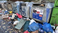 Vor Papiercontainern liegt eine große Menge Müll, z.B. Papier, blaue Säcke und Folien