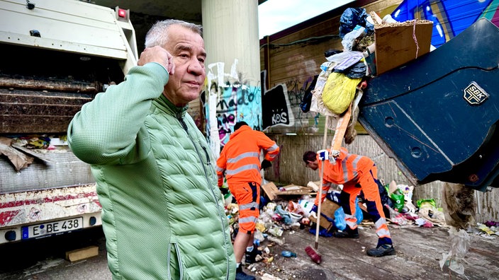 Ein Mann in grüner Jacke steht im Vordergrund, dahinter steht ein Müllwagen, daneben arbeiten zwei Mitarbeiter der Entsorgungsbetriebe in orangenen Jacken und heben Müll vom Boden auf. In der Hand haben sie einen Besen.