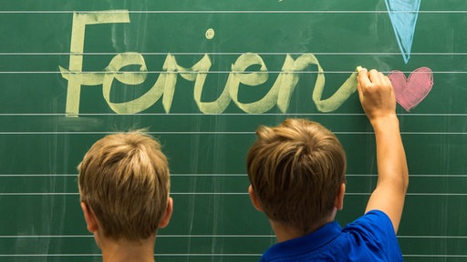 Zwei Kinder schreiben "Ferien" an eine Tafel