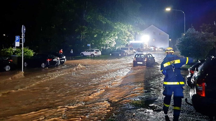 Eine Straße ist von braunem Wasser geflutete, in der Mitte steckt ein schwarzes Auto fest, rechts im Bild ein Mann in blau-gelber Uniform vom Technischen Hilfswerk