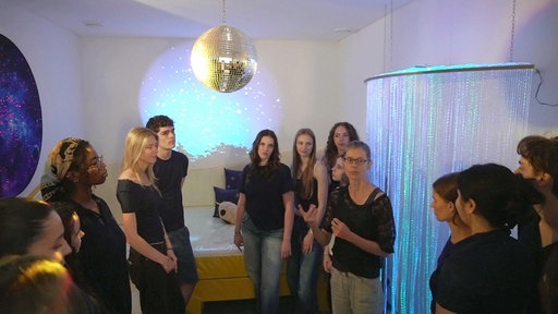 Jugendliche stehen gemeinsam in einem Raum, in dem eine Diskokugel hängt, ein Bett steht im Hintergrund.