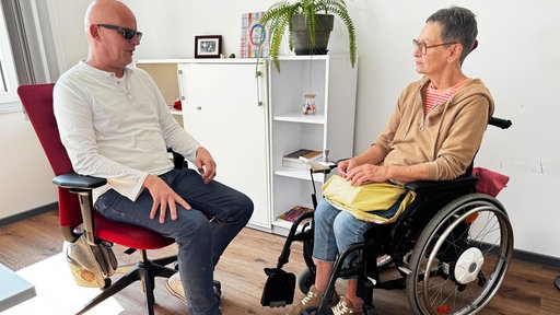 Ein blinder Berater mit dunkler Sonnenbrille sitzt einer Frau im Rollstuhl gegenüber
