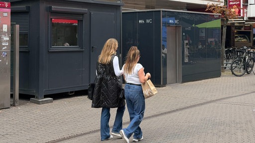 Zwei Frauen laufen an einer öffentlichen Toilette in der Innenstadt vorbei. 