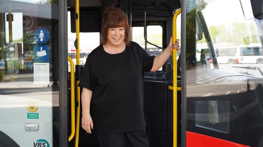 Eine lachende Frau mit rot-brauner Ponyfrisur steht an der offenen Tür eines Linienbusses. 