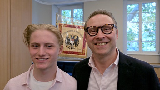 Constantin und Hubertus Dieckmann stehen vor zwei Fenstern und einer Fahne mit der Aufschrift "Männergesangverein" und lächeln in die Kamera