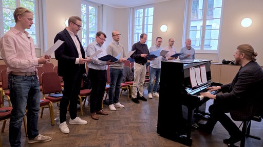 Einige Männer stehen in einem hellen Raum um ein Klavier herum und halten Notenblätter in der Hand