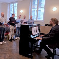 Einige Männer stehen in einem hellen Raum um ein Klavier herum und halten Notenblätter in der Hand