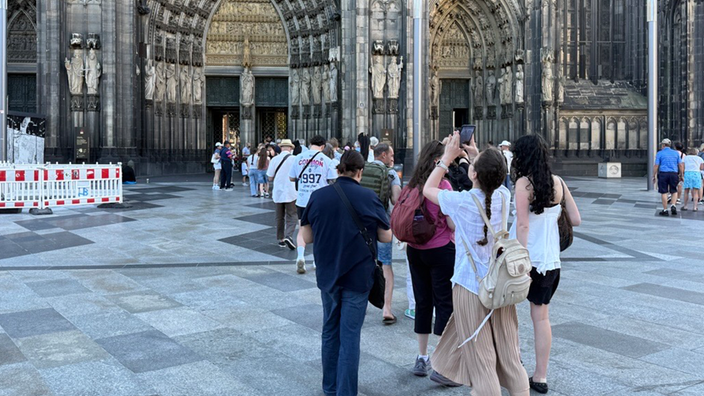 Dutzende Menschen stehen auf der Domplatte vor dem Hauptportal des Doms Schlange