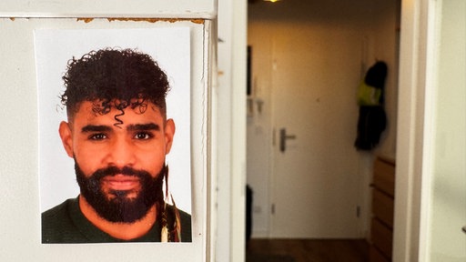 Ein kleines Foto von Pedro Corona an einem Regal in seiner Wohnung mit der Wohnungstür im Hintergrund 