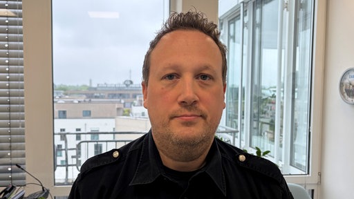 Ein Mann mit kurzen Haaren und dunkler Uniform steht vor dem Fenster in einem Büro und guckt freundlich in die Kamera