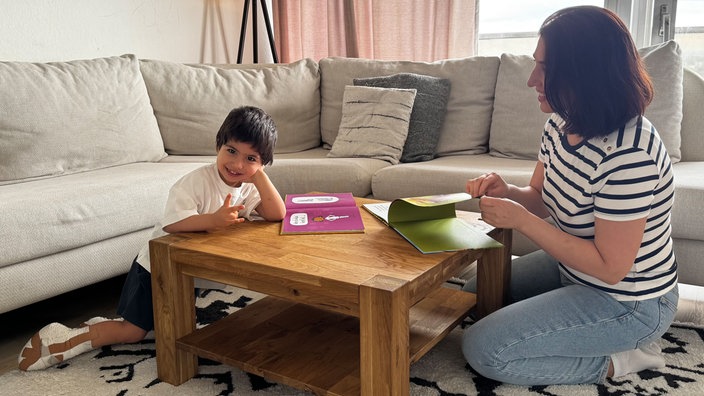 Der zweijährige Raybod sitzt am Couchtisch, lacht in die Kamera und zeigt zwei Finger. Seine Füße sind eingegipst. Neben ihm sitzt seine Mutter und blättert in einem Buch.
