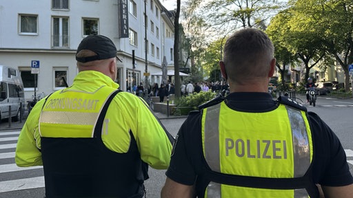 Ein Polizist und ein Mitarbeiter des Ordnungsamts von hinten fotografiert in gelber Warnweste vor der langen Schlange am Impi-Café