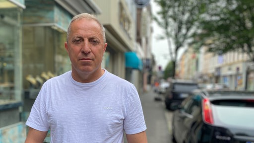 Mann im T-Shirt schaut ernst in die Kamera, im Hintergrund eine Straße mit Läden und parkenden Autos