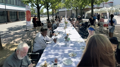 mehrere menschen sitzen an einer langen, gedeckten Tafel und unterhalten sich