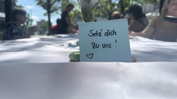 Auf einem Tisch steht ein blaues Kärtchen mit der Aufschrift: "Setz dich zu uns"