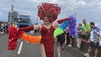 Eine Drag-Queen in rotem Glitzer-Abendkleid und roter, großer Kopfbedeckung, auf der "Pride" steht, läuft jubelnd in der Parade mit.