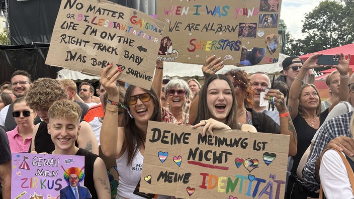 Weibliche Gäste stehen jubelnd am Paradenrand und halten selbstgemachte Demonstrationsschilder hoch.